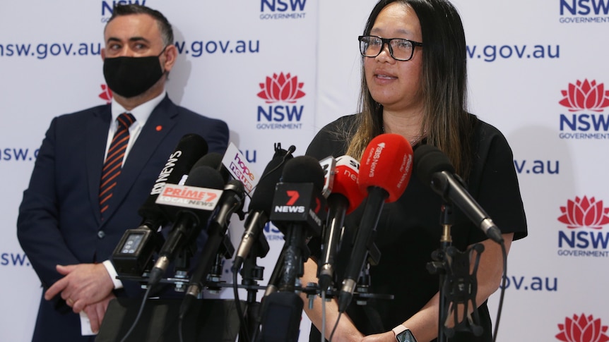 Dr Nhi Nguyen stands in front of a lectern covered with news microphones while NSW deputy premier John Barilaro looks on.
