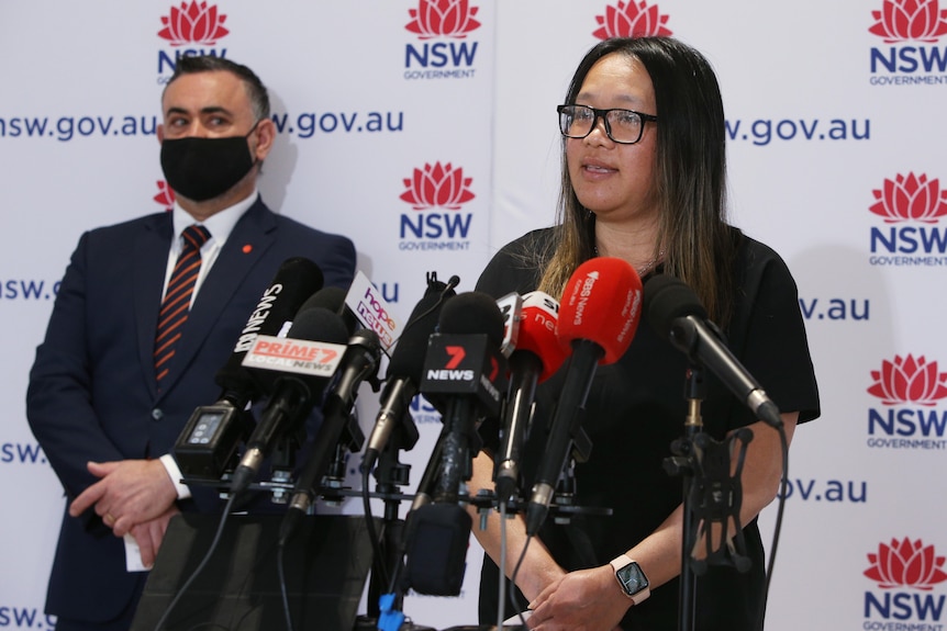 Dr Nhi Nguyen stands in front of a lectern covered with news microphones while NSW deputy premier John Barilaro looks on.
