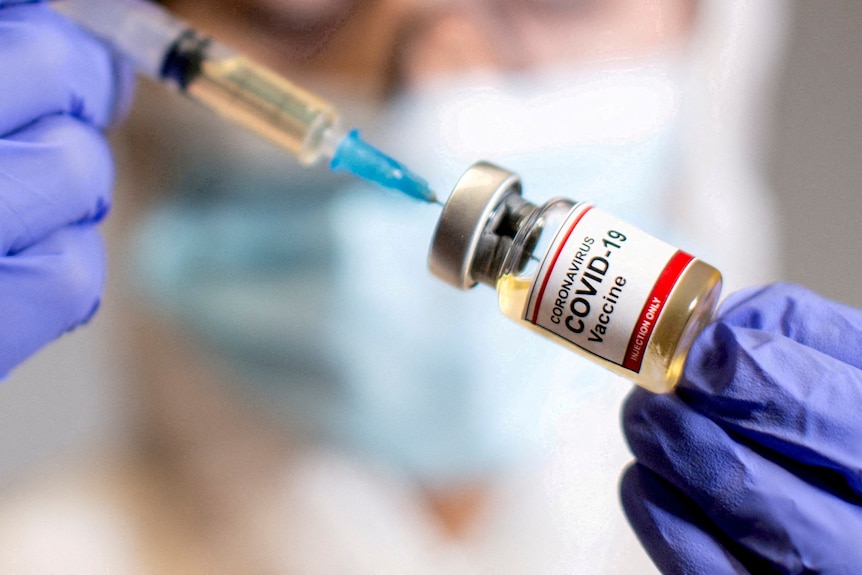 A woman holds a medical syringe and a small bottle labeled "Coronavirus COVID-19 vaccine" 