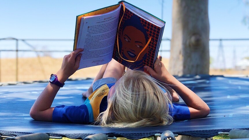 A young child lies on her back in an outside rural setting, reading a book.