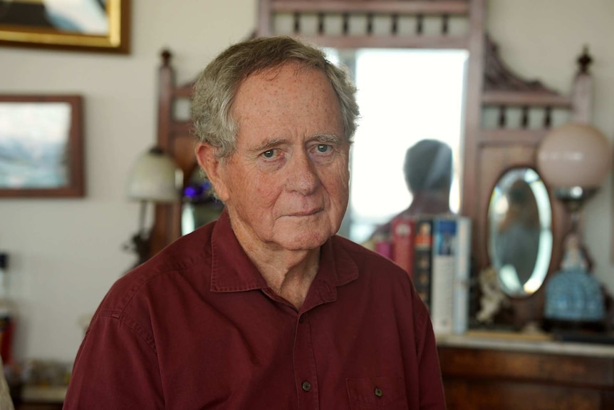 An older man sits in a living room with a grave expression on his face.