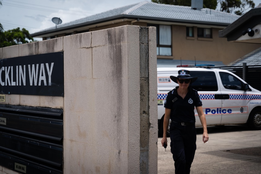 A female police officer walks toward the camera outside a unit complex with a police van parked