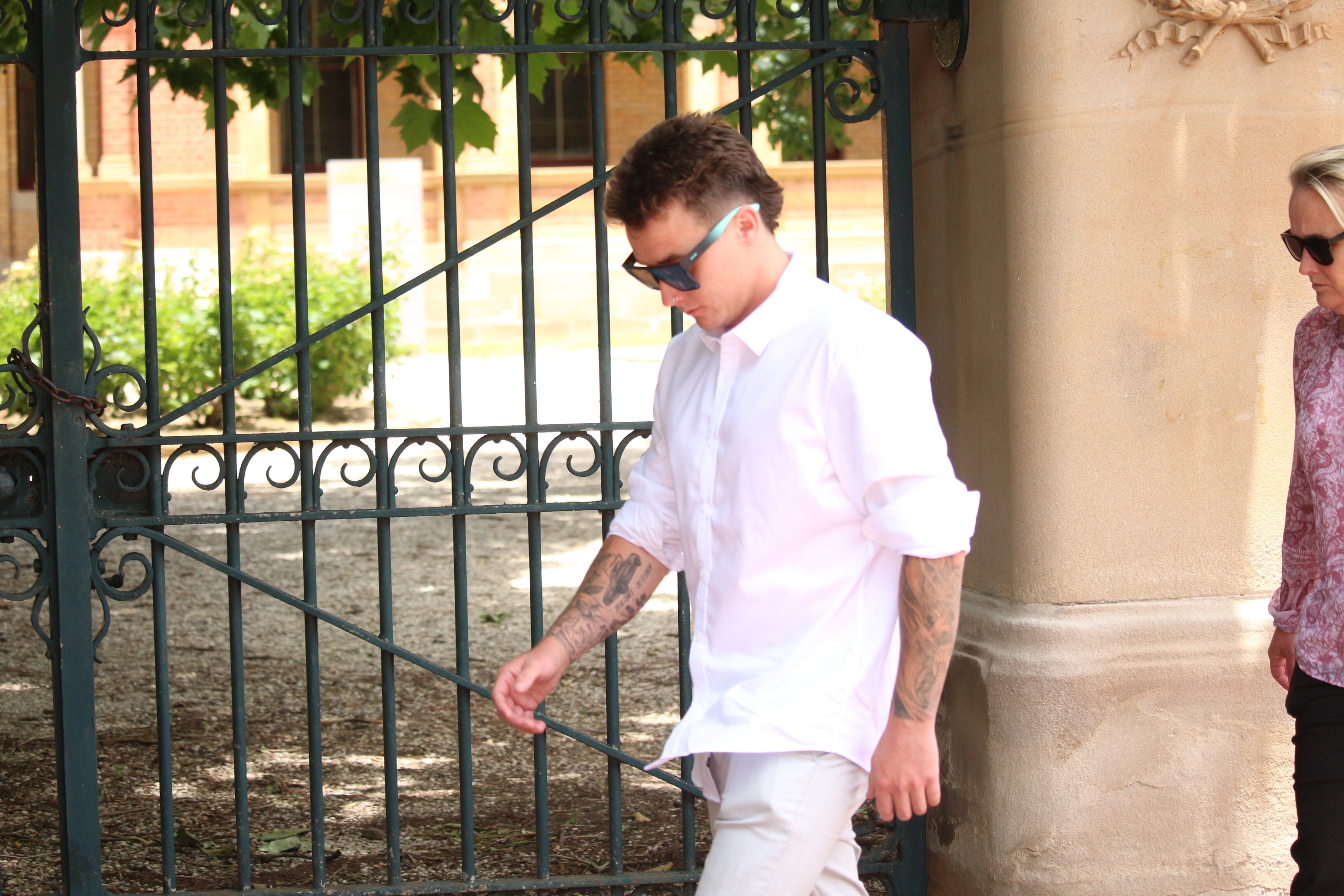 Un hombre con cabello rubio corto, gafas de sol y una camisa blanca con botones camina frente a una valla de hierro forjado.