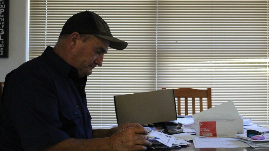 A man sits side-on to the camera looking through bills at a kitchen table