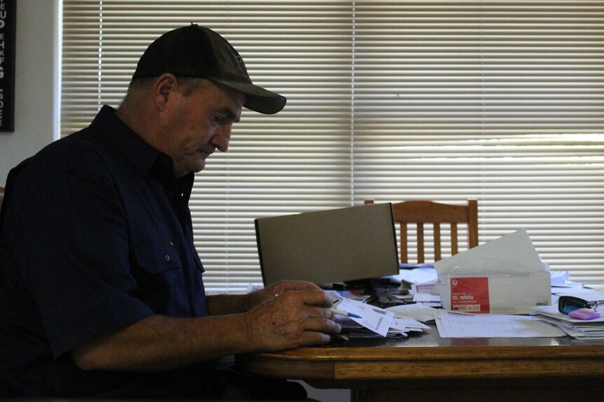 A man sits side-on to the camera looking through bills at a kitchen table