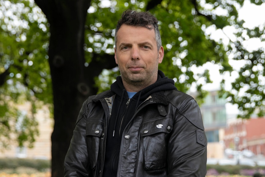 A man wearing a black leather jacket looks seriously at the camera.