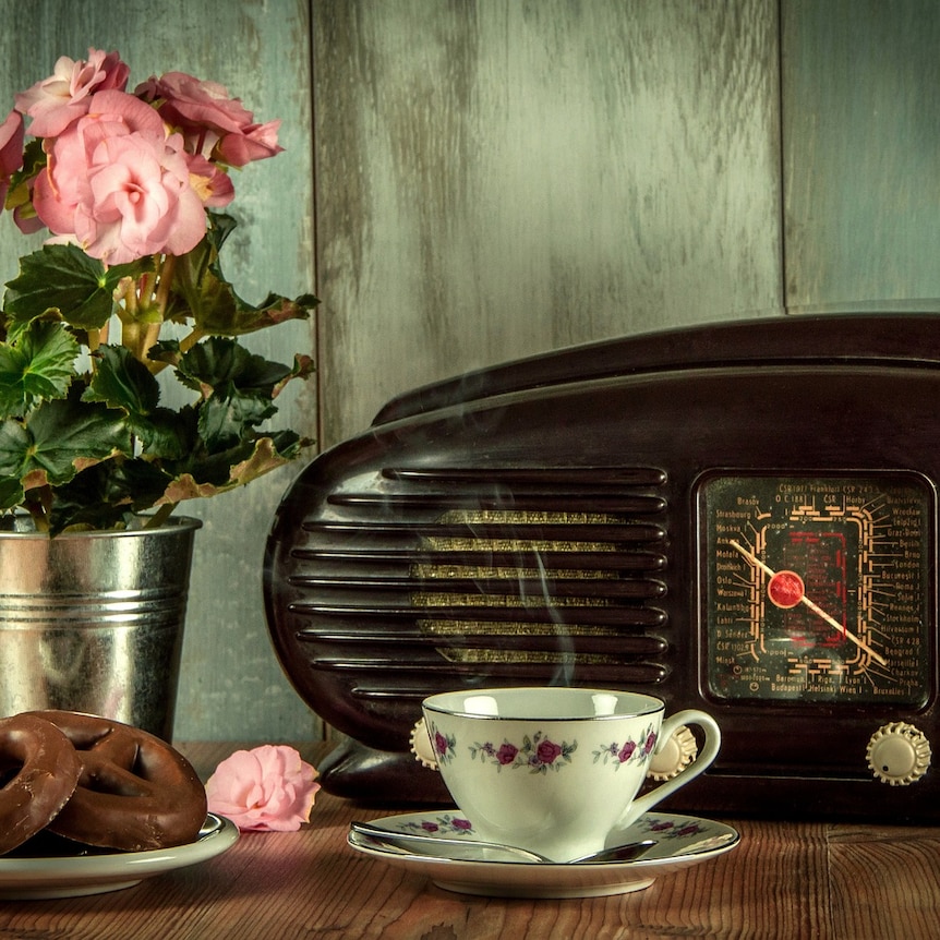 An old fashioned console radio sitting on a table with a cup of tea, plate of chocolate biscuits and vase of flowers around it.