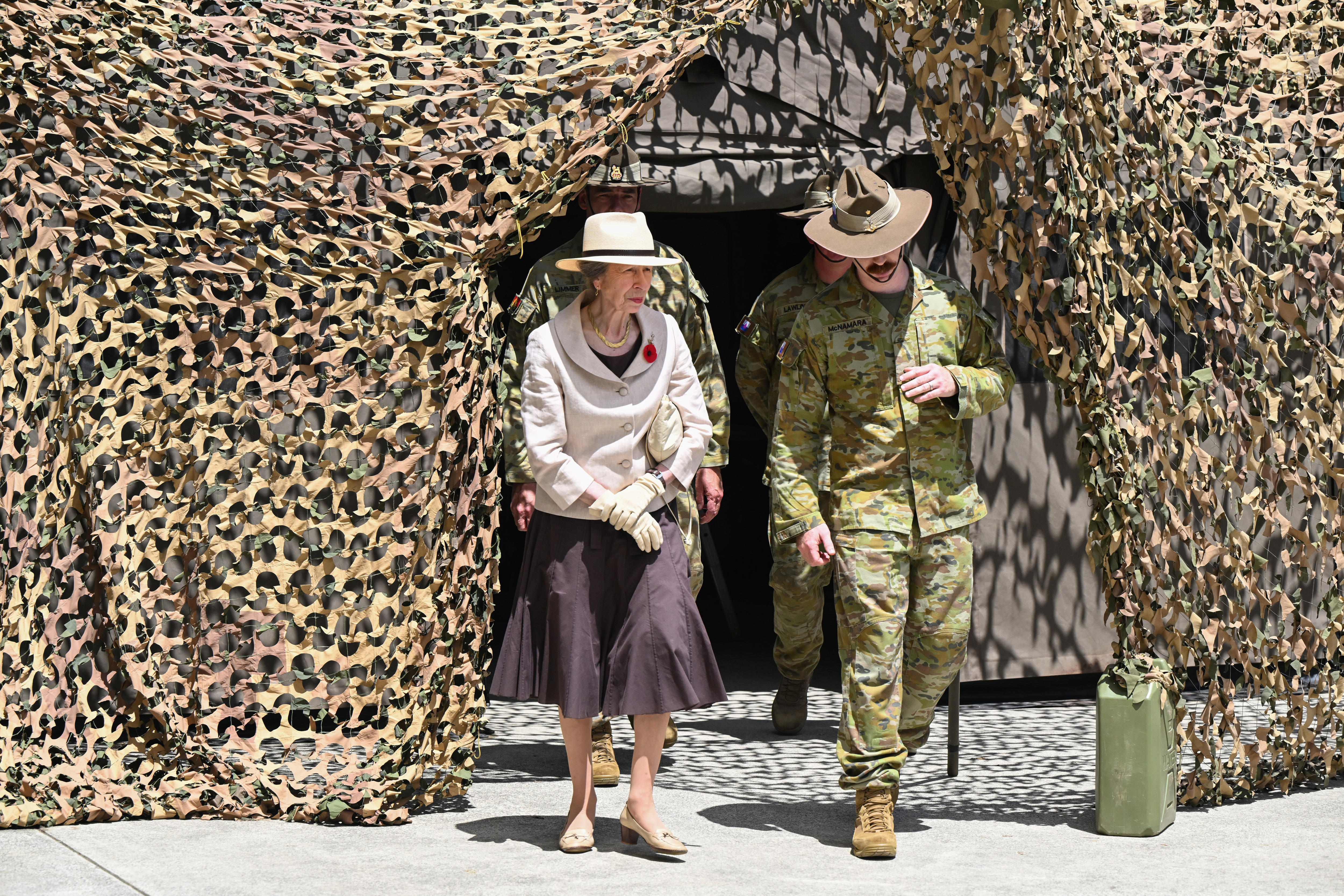Uma senhora idosa vestida com roupas formais caminha ao lado de um soldado uniformizado.