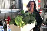 A woman is standing holding a box filled with green vegetables and looking at the camera
