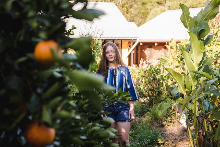 A teenage girl wearing a blue top and denim shorts stands next to a fruit tree.