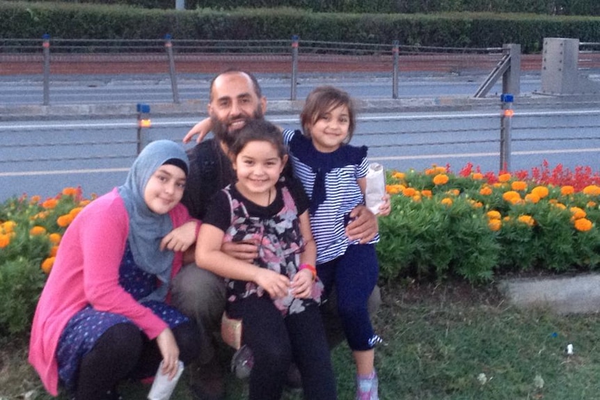 Older man smiles with three young girls aged under ten on side of road with flowers behind them.