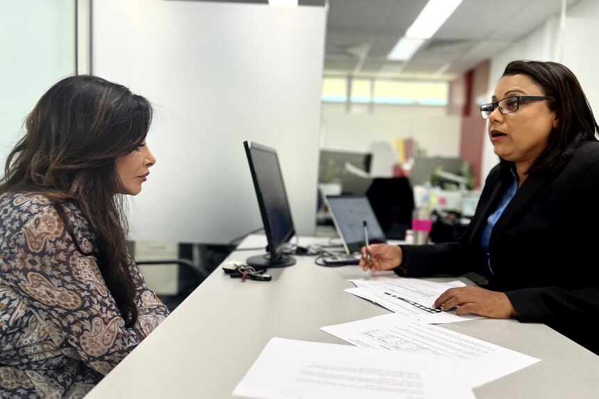 Two women sit across a table from each other, with papers and a computer screen in between.