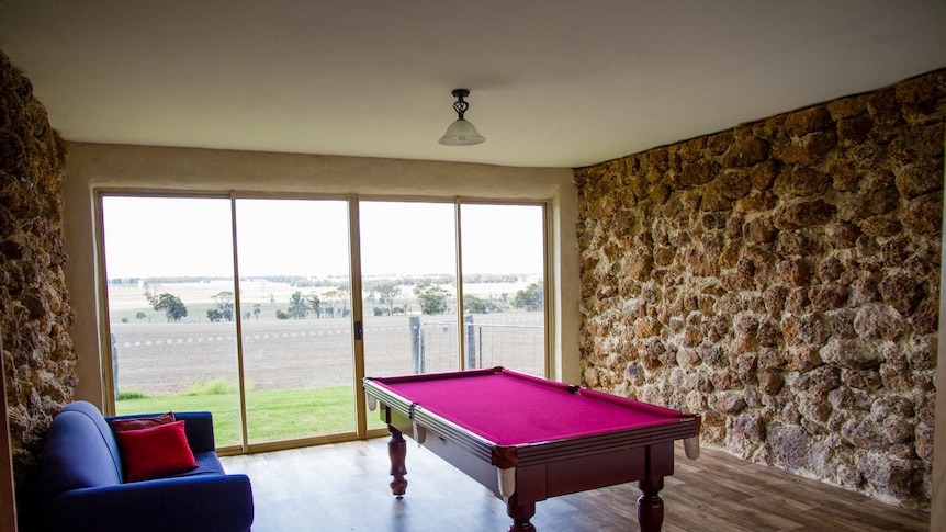 A room with a couch, pool table, view of a farm and two rock walls.
