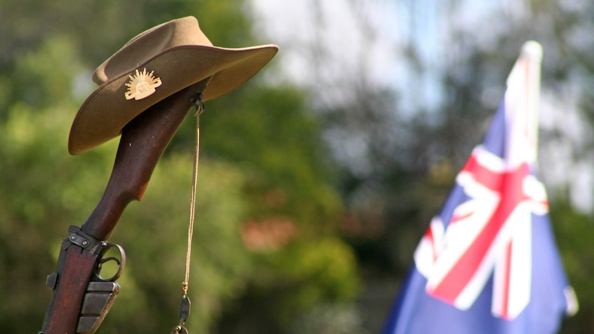 An old WWI rifle with an Anzac slouch hat on top with rising sun badge next to an Australian flag.