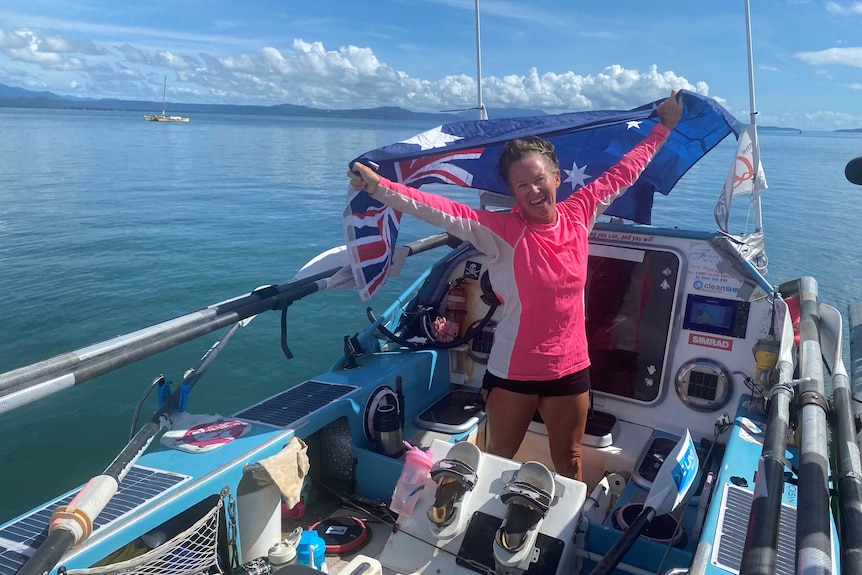 A woman on board a small rowboat packed with gear lifts an Australian flag.