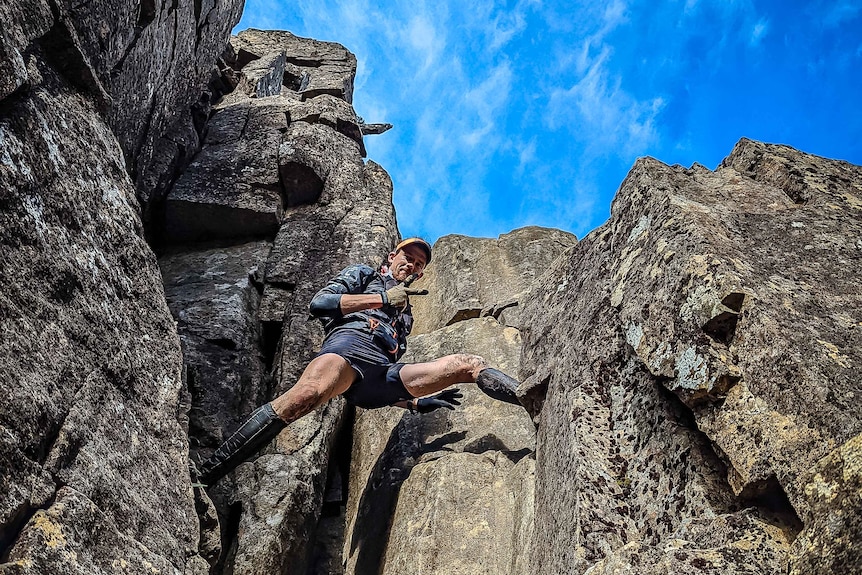 A man between vertical sections of rock on a mountain, looking down at the camera.