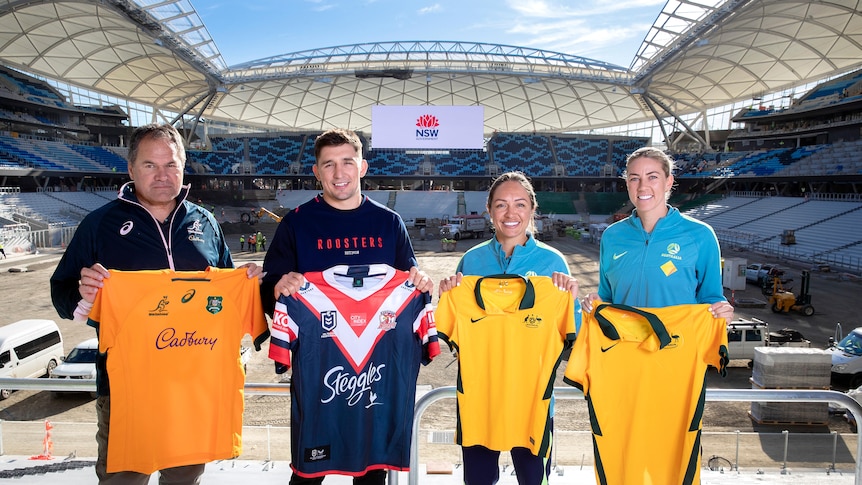 Two men and two women hold up sports shirts inside a stadium being renovated