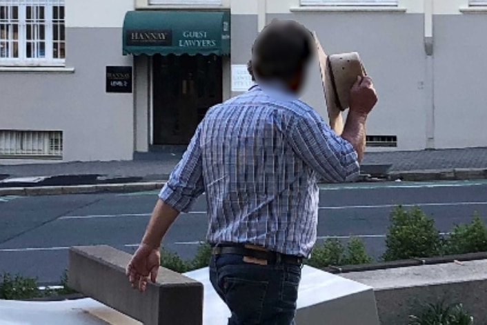 A man in jeans and checked shirt, walking away, holding a hat up against the side of his face