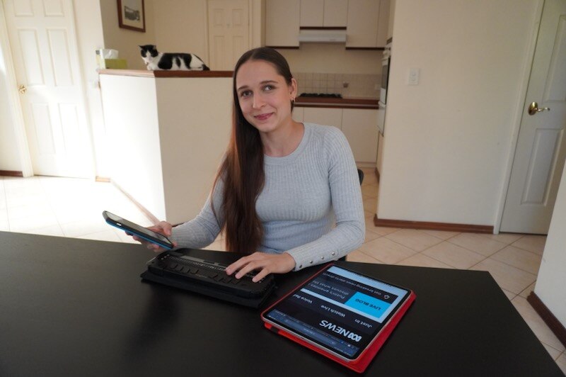 Vanessa, a young woman with long brown hair, sits at a table smiling, holding a phone with her Brailliant and an iPad.