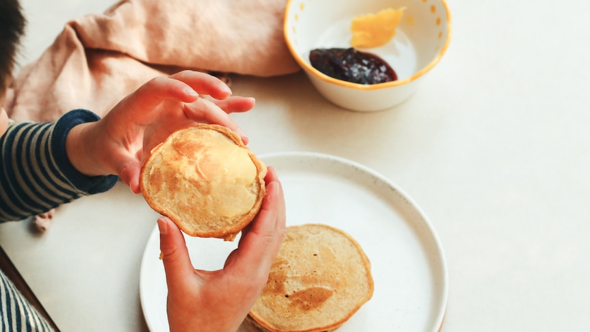 Child's hands hold a pikelet, with another pikelet on their plate. Jam and butter are in the background.