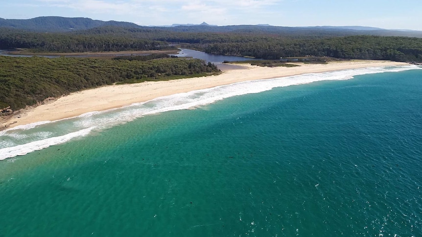 An aerial shot of a beach with turquoise water and white sand. Behind it are trees, an inlet, and a mountain.