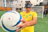 A male blind footballer wears a mask over his eyes and is holding a soccer ball out in front of him.