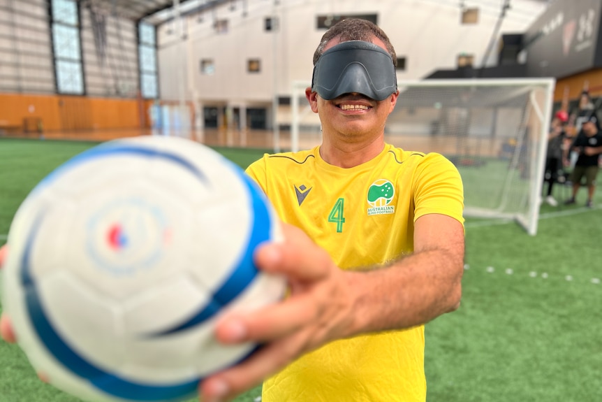 A male blind footballer wears a mask over his eyes and is holding a soccer ball out in front of him.