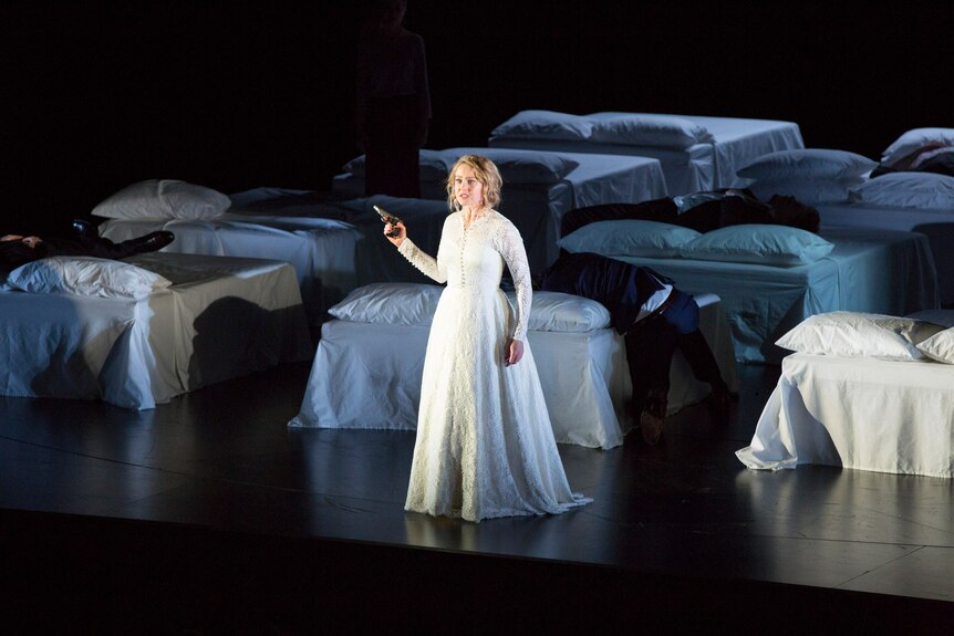 White woman wears floor-length white dress and stands on stage holding handgun among white linen beds.