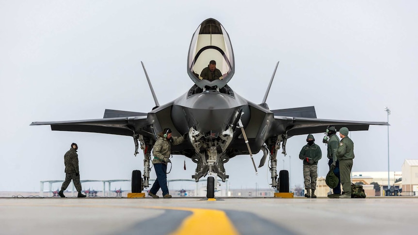 Laying on the tarmac, the viewer looks up to a F-35 jet parked as military workers attend to landing procedures.