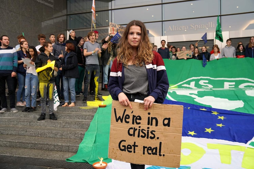 Luisa Neubauer, standing in front of other activists, holds a sign saying "We're in a crisis. Get real".