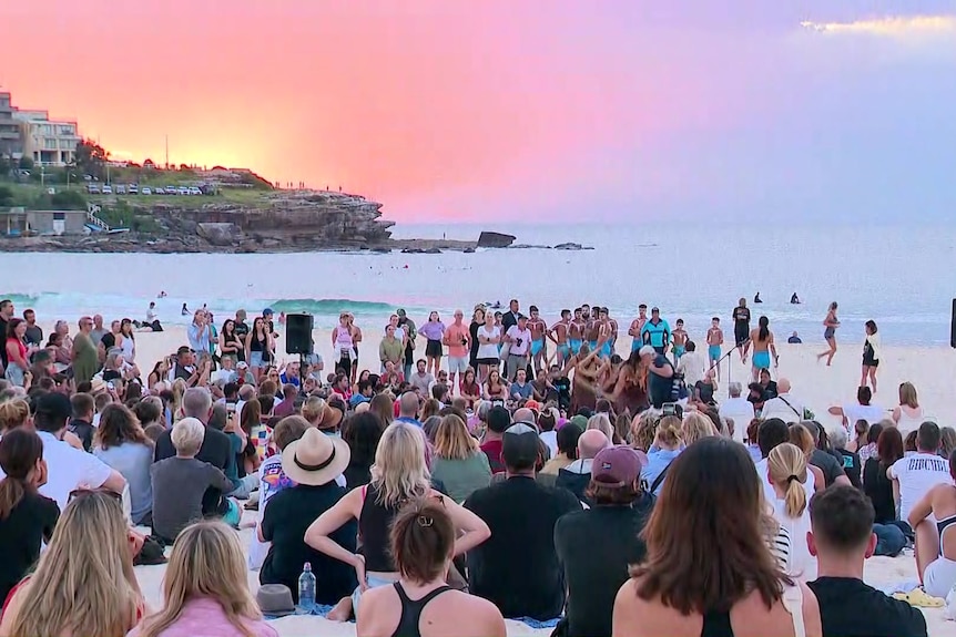 Crowds gather on the sand in front of the water under a yellow, orange, pink, purple and blue dawn sky.