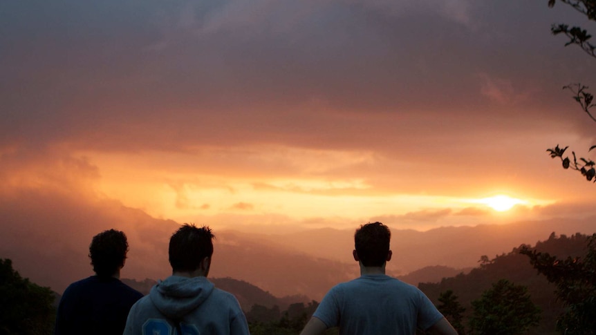 From behind, you see the silhouettes of three men looking out to mountaintops bathed in an orange glow at sunset.