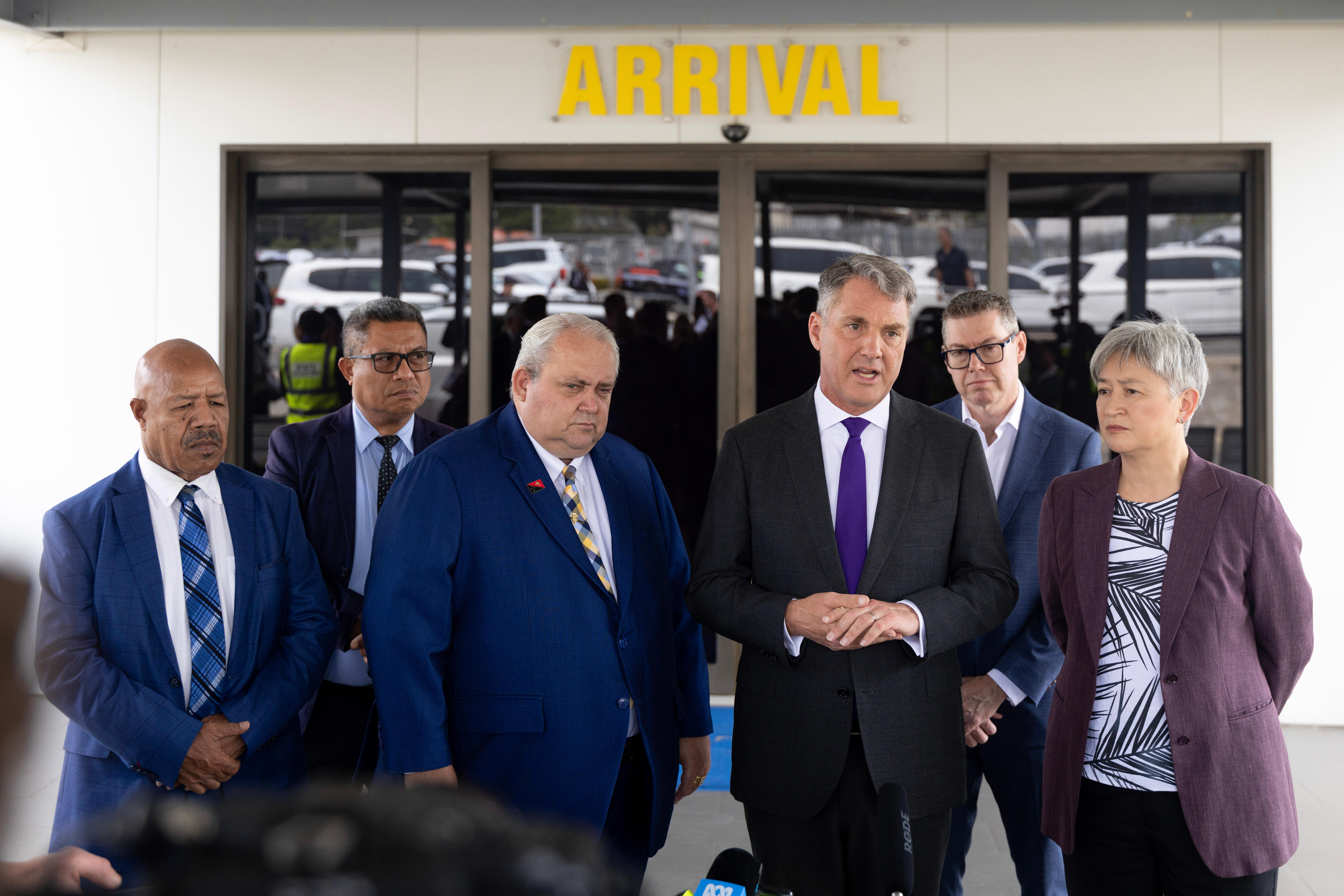 Five men in black or blue suits and a woman in a purple suit jacket in front of a sign saying 'arrival'.