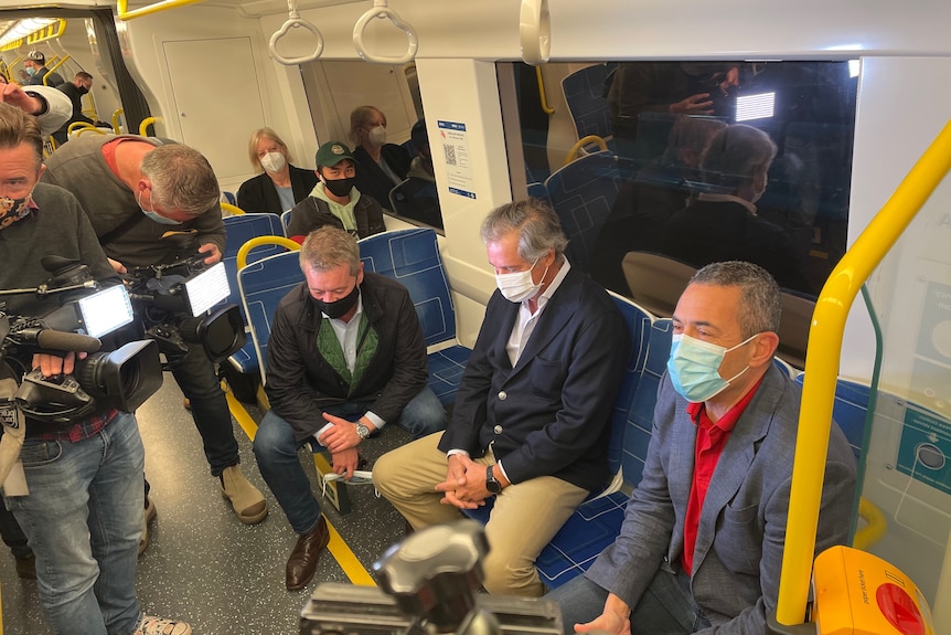 A man sits on a train wearing a face mask, but his eyes are smiling, surrounded by news cameras.