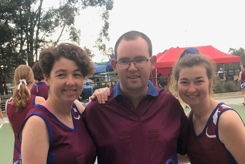 Damian stands in between two female netballers all of them wear maroon and navy team colours with marquees in the background