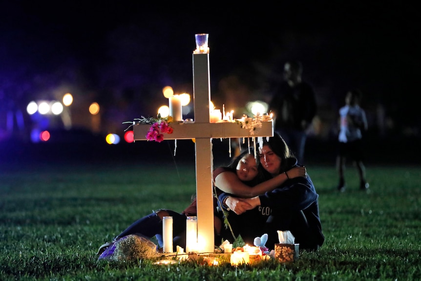 Two young women sit at night crying and hugging in front of a large white cross covered in candles. 