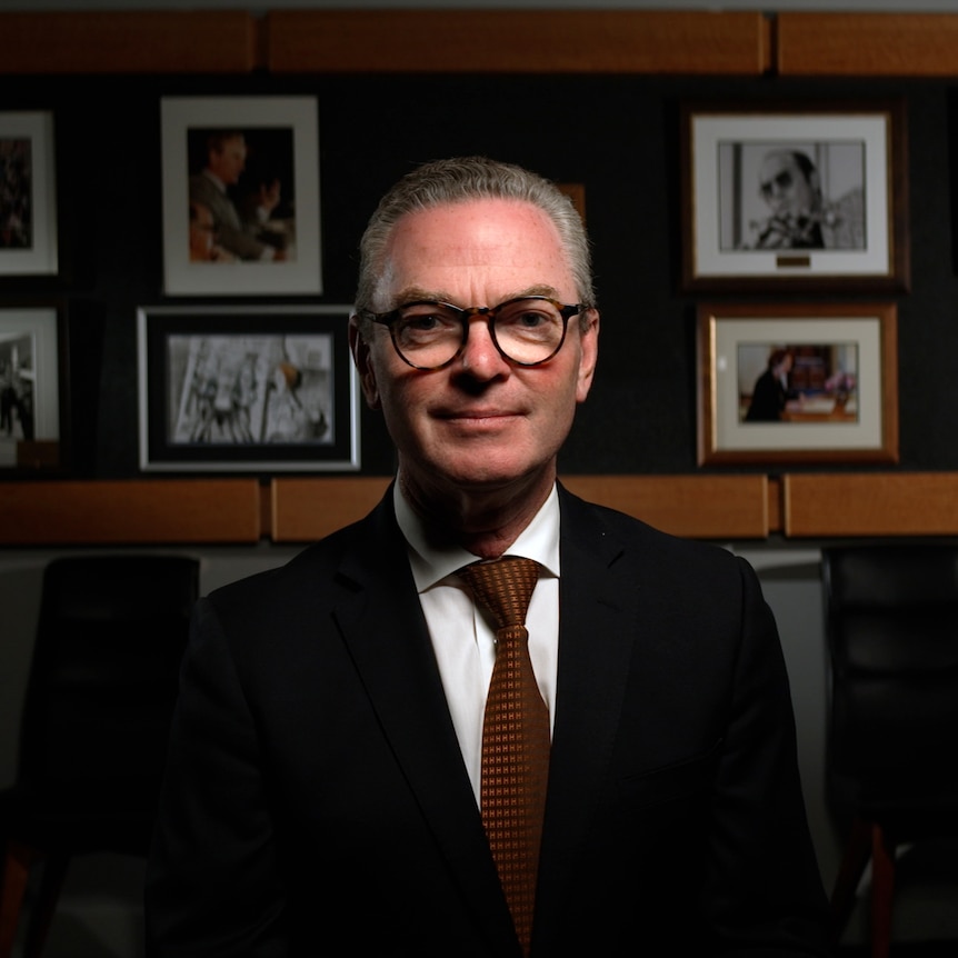 Dressed in a dark suit and tie, Christopher Pyne sits in a room, looking into camera with a neutral expression. 