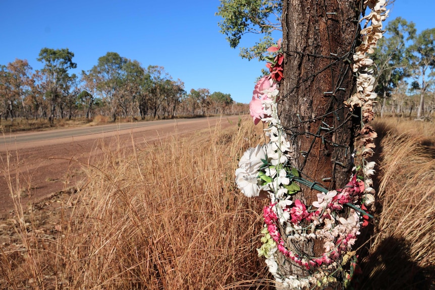 Pink and white flowers and fairy lights are tied to a tree, next to long yellow grass by the highway