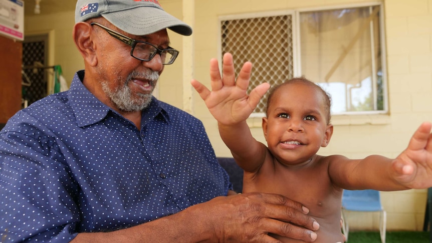 Older Torres Strait Islander man wearing a cap and glasses smiles at his granddaughter who is reaching for the camera.