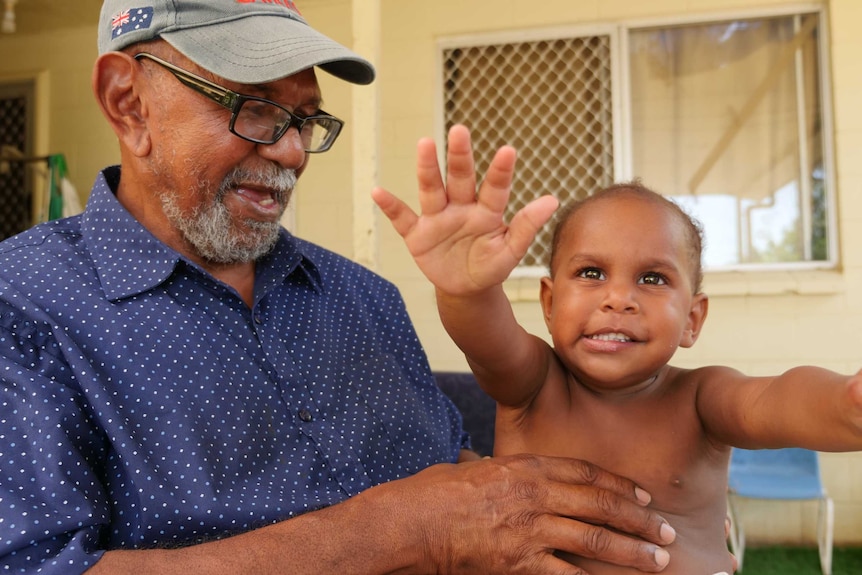 Older Torres Strait Islander man wearing a cap and glasses smiles at his granddaughter who is reaching for the camera.