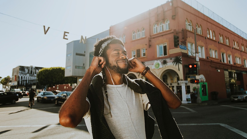 A photo of a man on the street holding headphones into his ears and smiling with his eyes closed.
