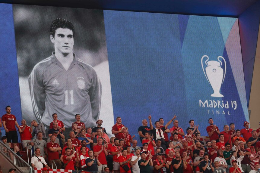 Fans in red stand in the grandstand with a big poster of a footballer with number 11 on his shirt.