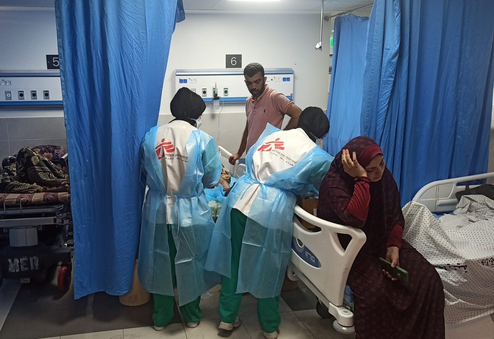 Two MSF workers are assisting a patient in a hospital room. A woman wearing a hijab sits on the bed, looking upset.