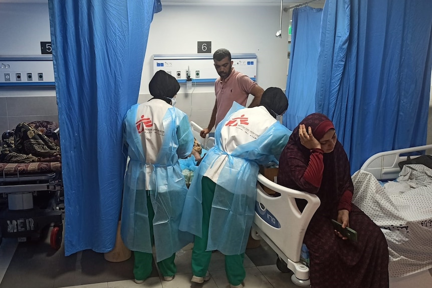 Two MSF workers are assisting a patient in a hospital room. A woman wearing a hijab sits on the bed, looking upset.