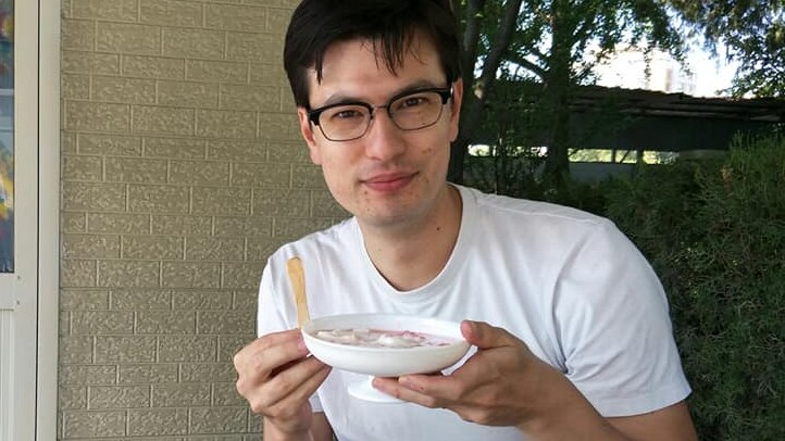 A man in a white t-shirt sits at a table outside holding a bowl of yoghurt and a chip.