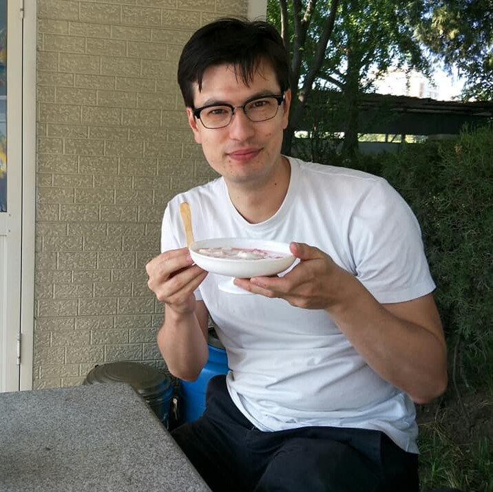 A man in a white t-shirt sits at a table outside holding a bowl of yoghurt and a chip.