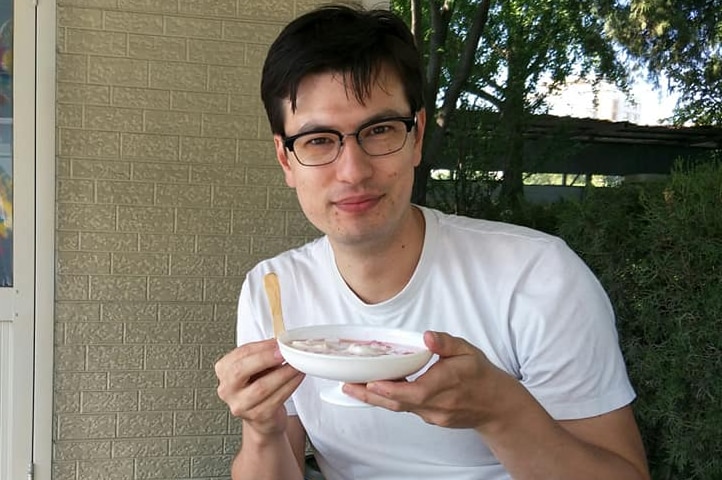 A man in a white t-shirt sits at a table outside holding a bowl of yoghurt and a chip.