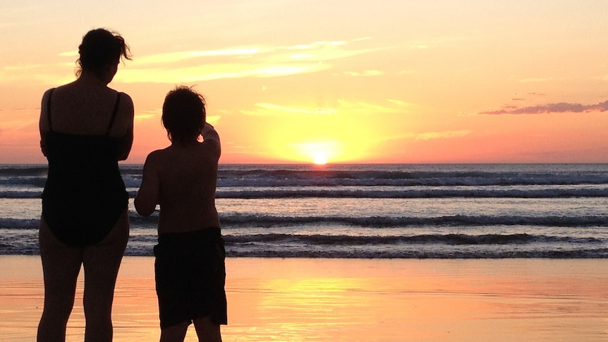 A mother and son standing together on a beach watching the sun set.