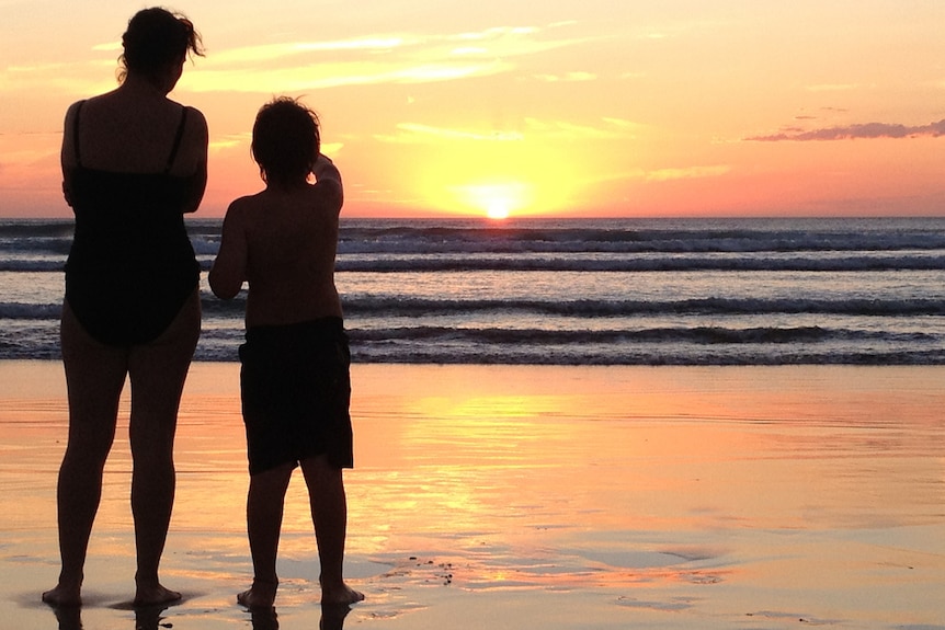 A mother and son standing together on a beach watching the sun set.