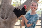 Young woman in a glittery gown, stands outside under a tree holding a black chicken. She is smiling.
